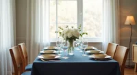 Navy tablecloth set with cream dinnerware, gold-rimmed glasses, and white flowers in a clear vase on a wooden dining table.