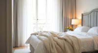 Cream fluffy blanket folded at the foot of a bed with white duvet and gray headboard.