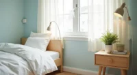 Pale blue walls frame a desk corner with white bedding, wooden nightstand, potted plant, and jute rug edge in natural window
