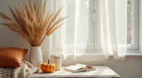 Cream tablecloth with rust pillows, knit blanket, wheat stalks, and ceramic pumpkin arranged on weathered wood.