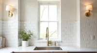 Two brass sconces flank a small window above a stainless steel sink with white subway tiles and marble countertop.